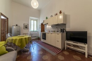Une cuisine compacte avec des meubles blancs, un four encastré, un coin repas avec une table et des chaises, et un téléviseur posé sur un meuble.