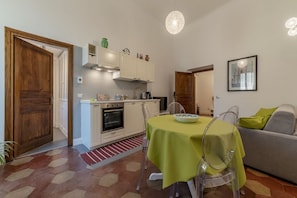 Une cuisine de taille réduite avec des meubles blancs, une table à manger recouverte d’une nappe verte et un canapé gris dans l’espace salon.