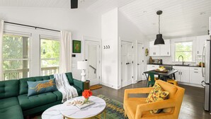 A spacious living room with a green sofa, a yellow armchair, and a marble coffee table. A kitchen is visible in the background with white cabinets and a black hanging light fixture. - Treefort (Atlanta)