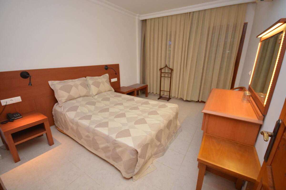 Habitación de hotel con cama, mesitas de noche, cómoda, espejo y ventana con cortinas.