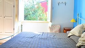 Un dormitorio con cama, ventana, silla, mesita de noche y lámpara.