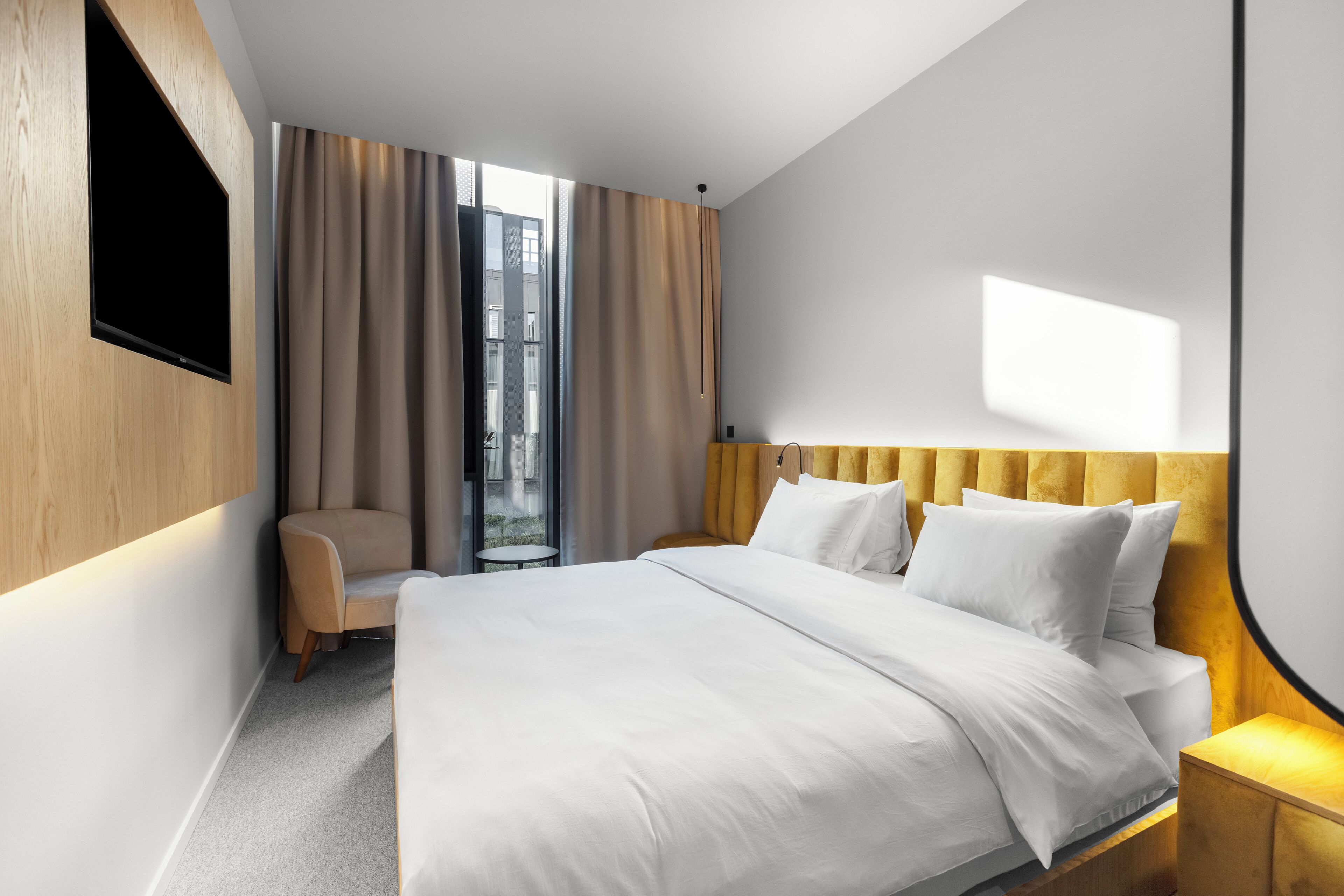 Habitación de hotel moderna con una cama grande, un televisor de pantalla plana montado en la pared, un sillón cómodo y vistas a la ciudad desde la ventana.