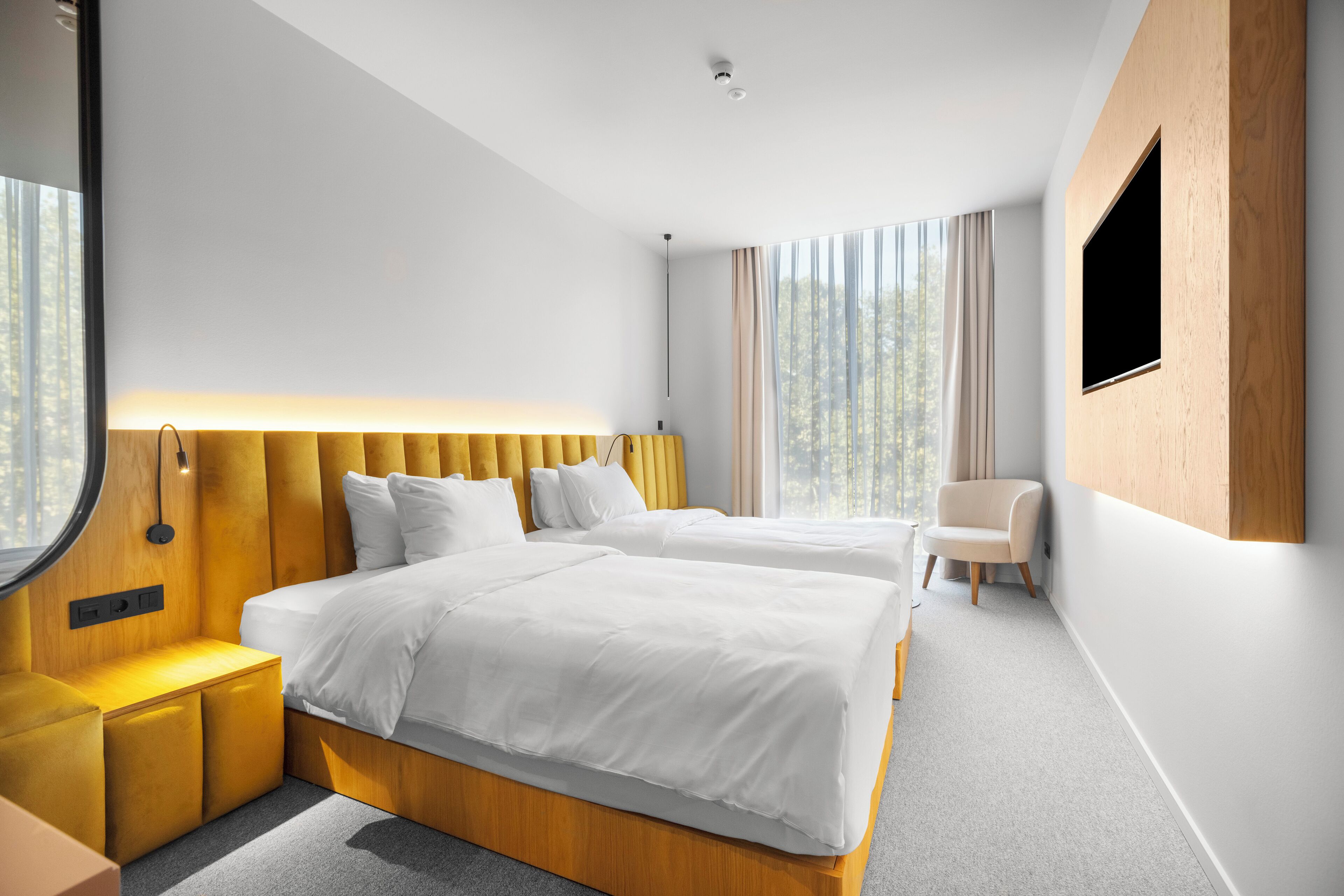 A modern hotel room with two beds, a wooden headboard, a bedside table, a chair, and a large window with curtains.
