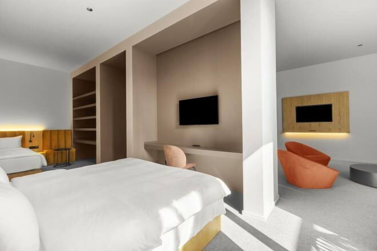 A modern hotel room with a bed, a TV mounted on the wall, a seating area with a chair, and a wooden panel on the wall.