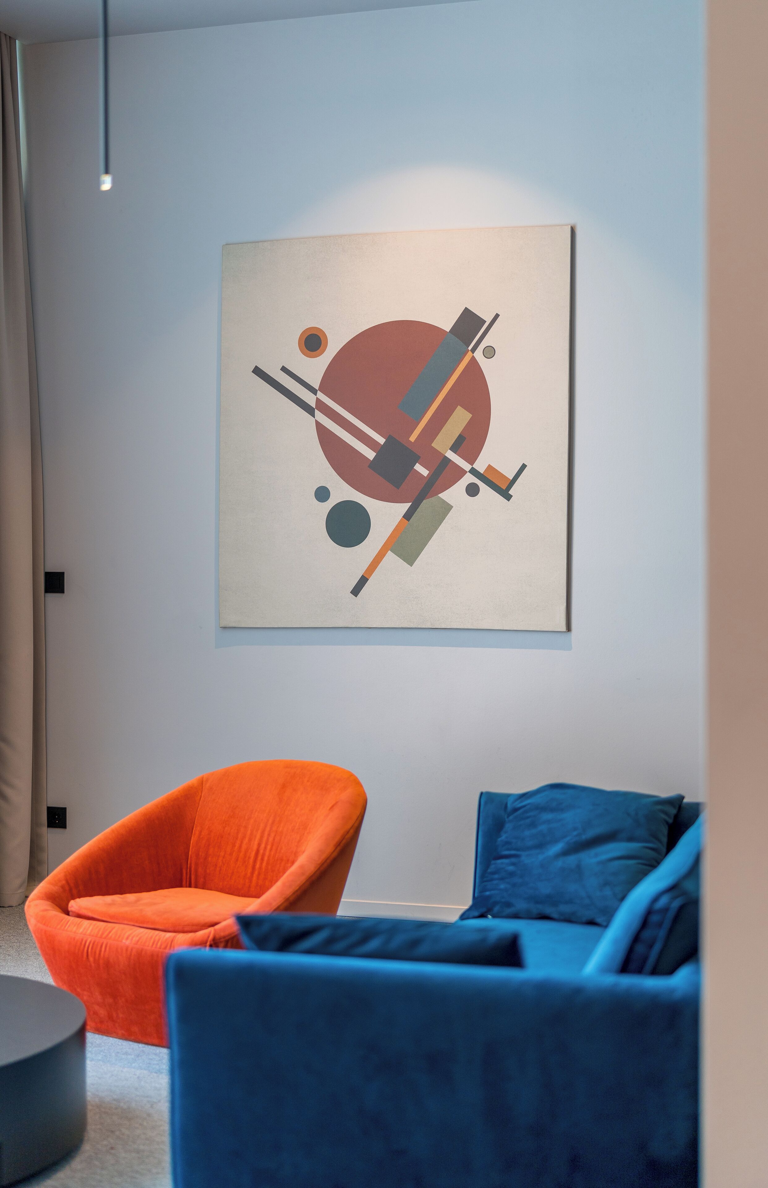 Una habitación moderna con un diseño minimalista, que presenta un cuadro abstracto grande en la pared, un sofá azul y una silla naranja.