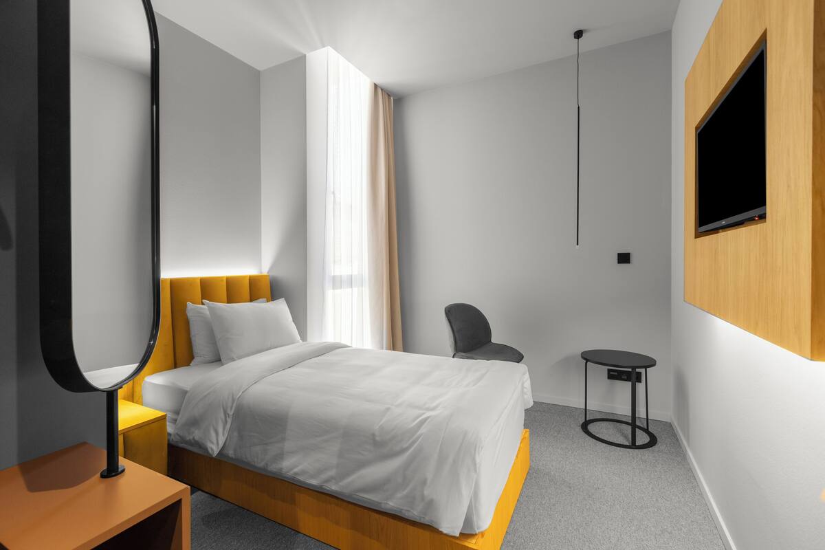 A modern hotel room with a bed, a bedside table, a chair, a small table, and a TV mounted on the wall.