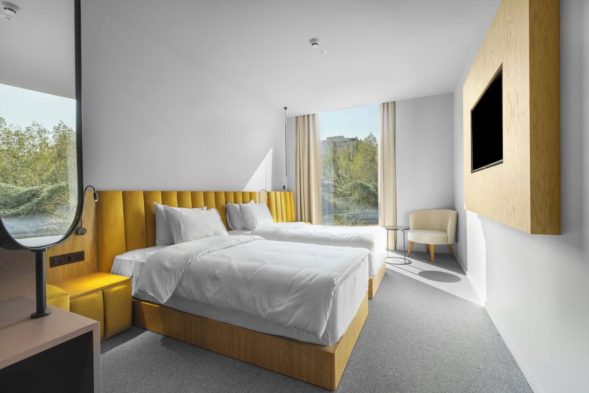 A modern hotel room with two beds, a small seating area, a mirror, and a view of trees.