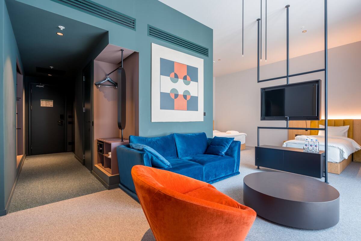A modern hotel room with a blue sofa, an orange armchair, a flat-screen TV, and a bed with a wooden headboard.