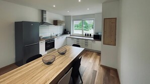 A modern kitchen with a wooden dining table, two black wire baskets, a refrigerator, a stove, and a window with a view of greenery. - 3-room gîte with terrace (Wintzenheim)