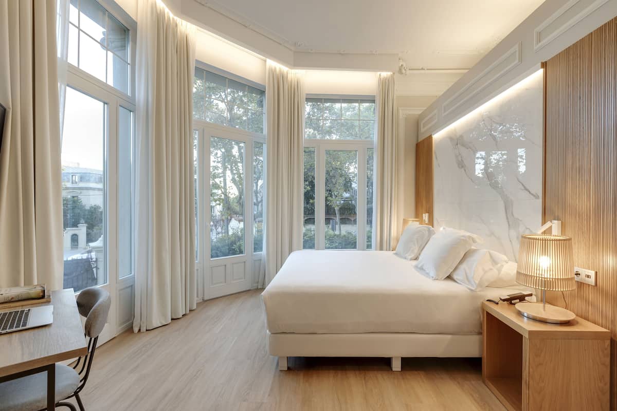 A bedroom with a large bed, a desk with a laptop, and a nightstand with a lamp.