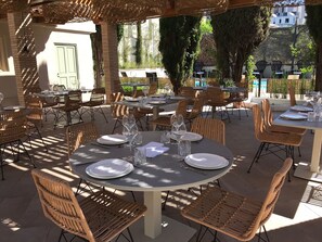 Un área de comedor al aire libre con mesas redondas y sillas de mimbre, rodeada de follaje y una piscina.