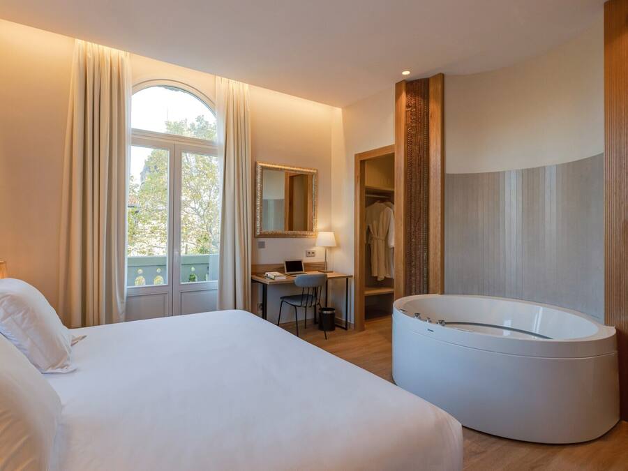 Una habitación de hotel con una cama grande, un escritorio con una computadora portátil, una bañera redonda y vistas al exterior.