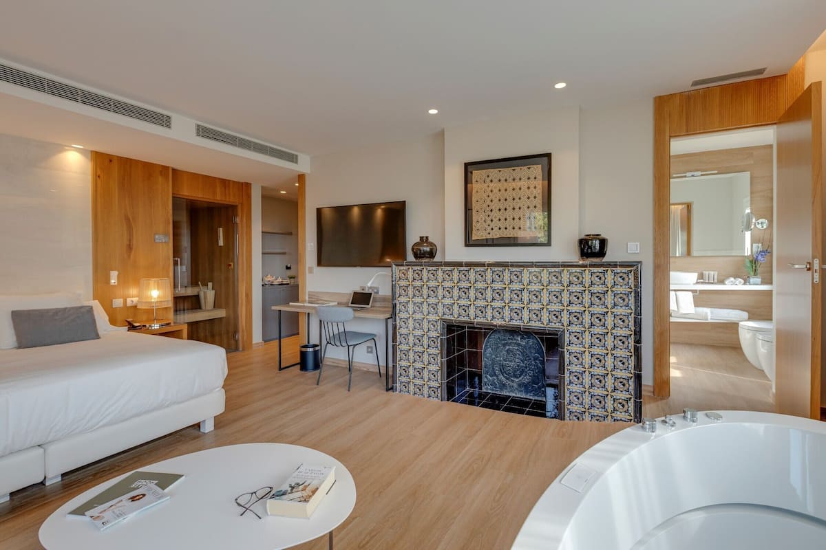 A modern hotel room with a bed, a desk, a fireplace, a bathroom, and a hot tub.