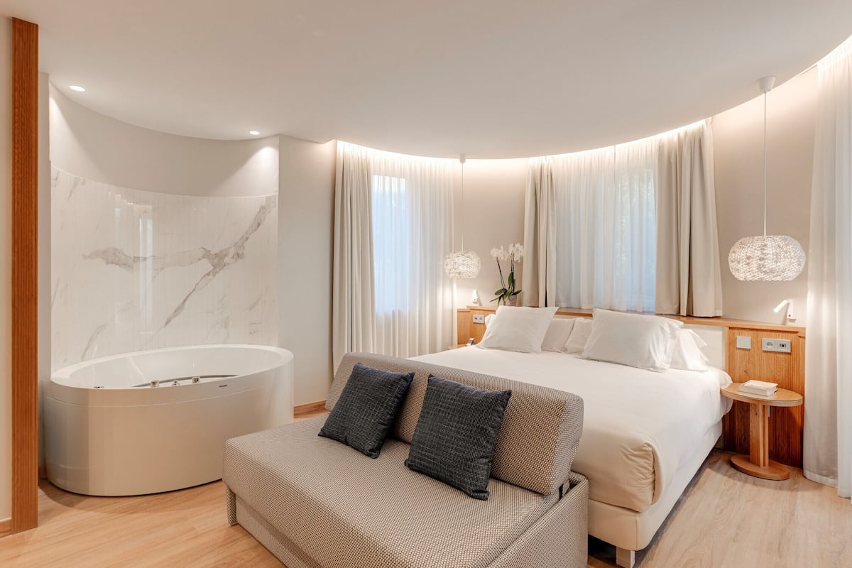 A modern hotel room with a curved ceiling, a large bed, a built-in bathtub, and a seating area.
