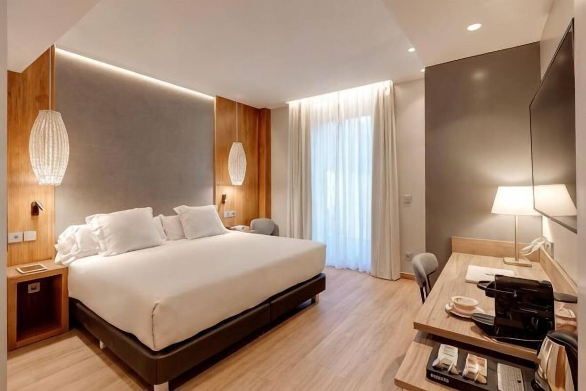 A modern hotel room with a large bed, bedside tables, a desk, a chair, and a large window with curtains.