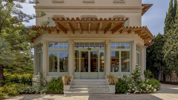 A two-story building with a traditional architectural style, featuring a wooden pergola over the entrance and a stone-paved courtyard.