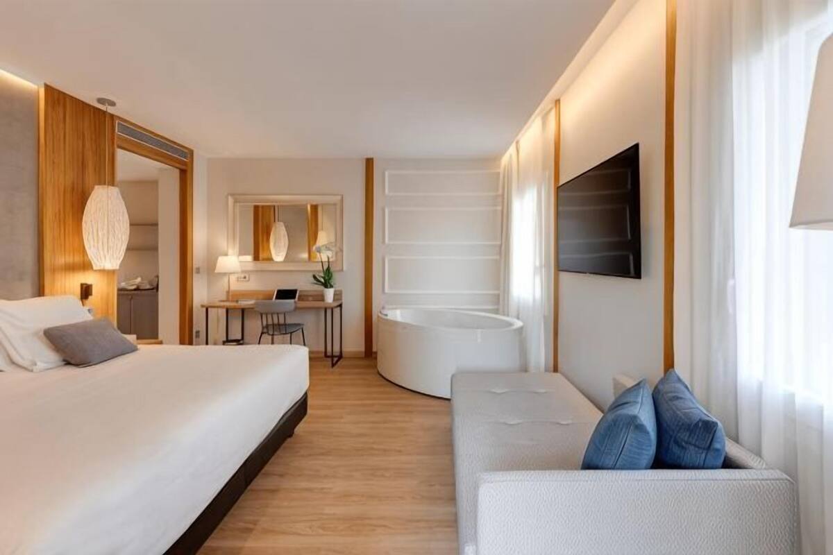 A modern hotel room with a bed, a sofa, a desk, a chair, a mirror, and a large window.