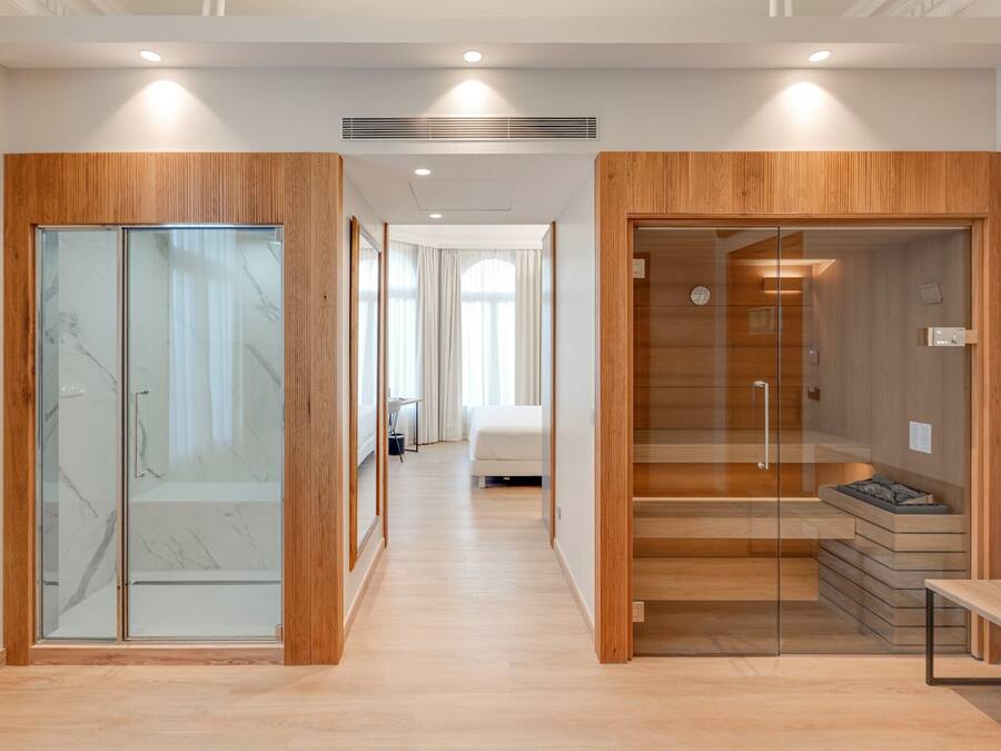 Ein modernes Hotelzimmer mit Holzboden, einer Duschkabine aus Glas und einem sichtbaren Schlafbereich mit Bett und Vorhängen.