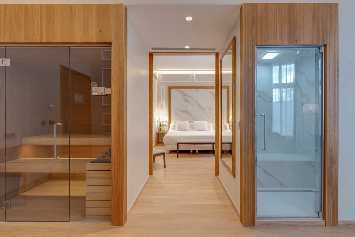 A modern hotel room with a wooden floor, a bed with white linens, a glass door, and a staircase leading to an upper level.
