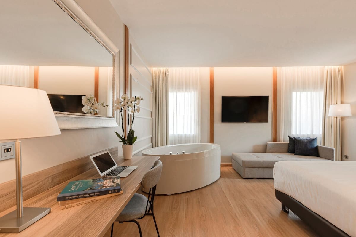 A modern hotel room with a desk, laptop, books, a large bed, a sofa, a bathtub, and a TV.