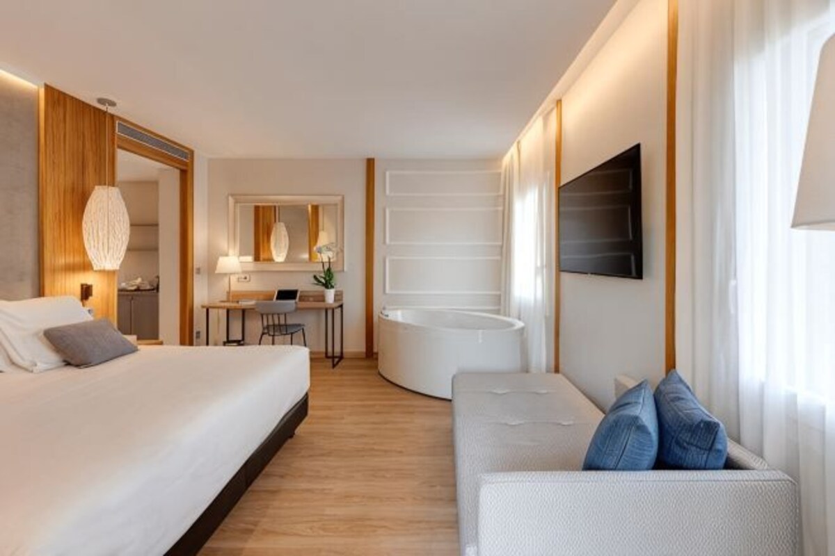 A modern hotel room with a bed, a sofa, a desk, a chair, a mirror, and a large window.