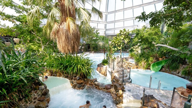 Een overdekt zwembadgedeelte met diverse waterattracties, waaronder een bubbelbad en een glijbaan, omringd door weelderige beplanting en rotsen.