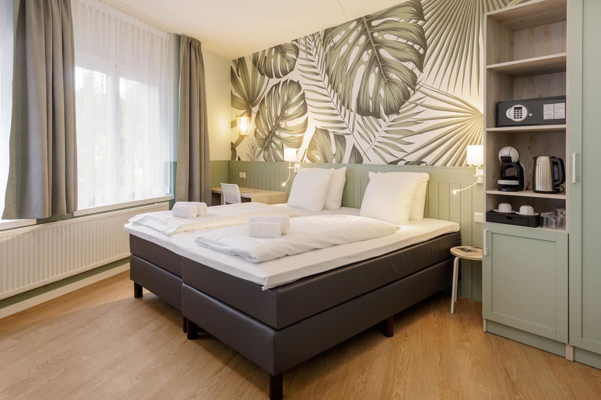 Une chambre d’hôtel moderne avec un grand lit, une tête de lit en bois, une table de chevet, une lampe, une étagère avec des articles de toilette et une fenêtre avec des rideaux.