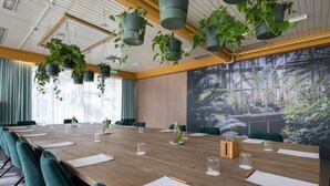 Een vergaderruimte met een lange houten tafel, groene stoelen, hangende plantenbakken en een groot scherm waarop een boslandschap wordt weergegeven.