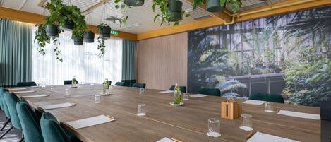 Uma sala de conferências com uma mesa de madeira comprida, cadeiras verdes, vasos suspensos e uma grande tela exibindo uma cena de floresta.