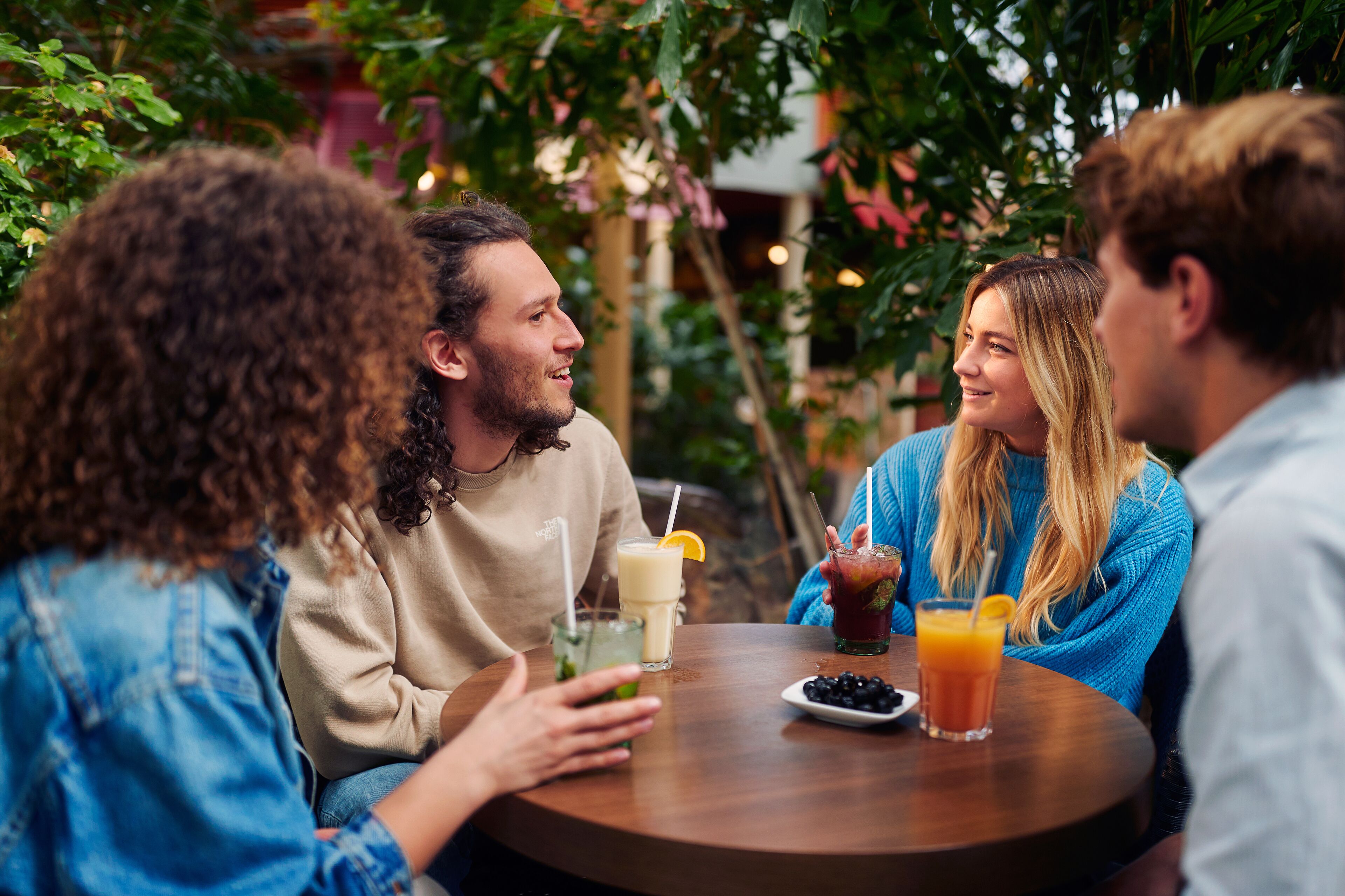 A group of people seated around a round wooden table with drinks, in an outdoor setting with greenery.
