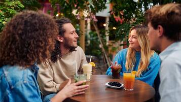 Een groep mensen zit rond een ronde houten tafel met drankjes, in een groene buitenomgeving.
