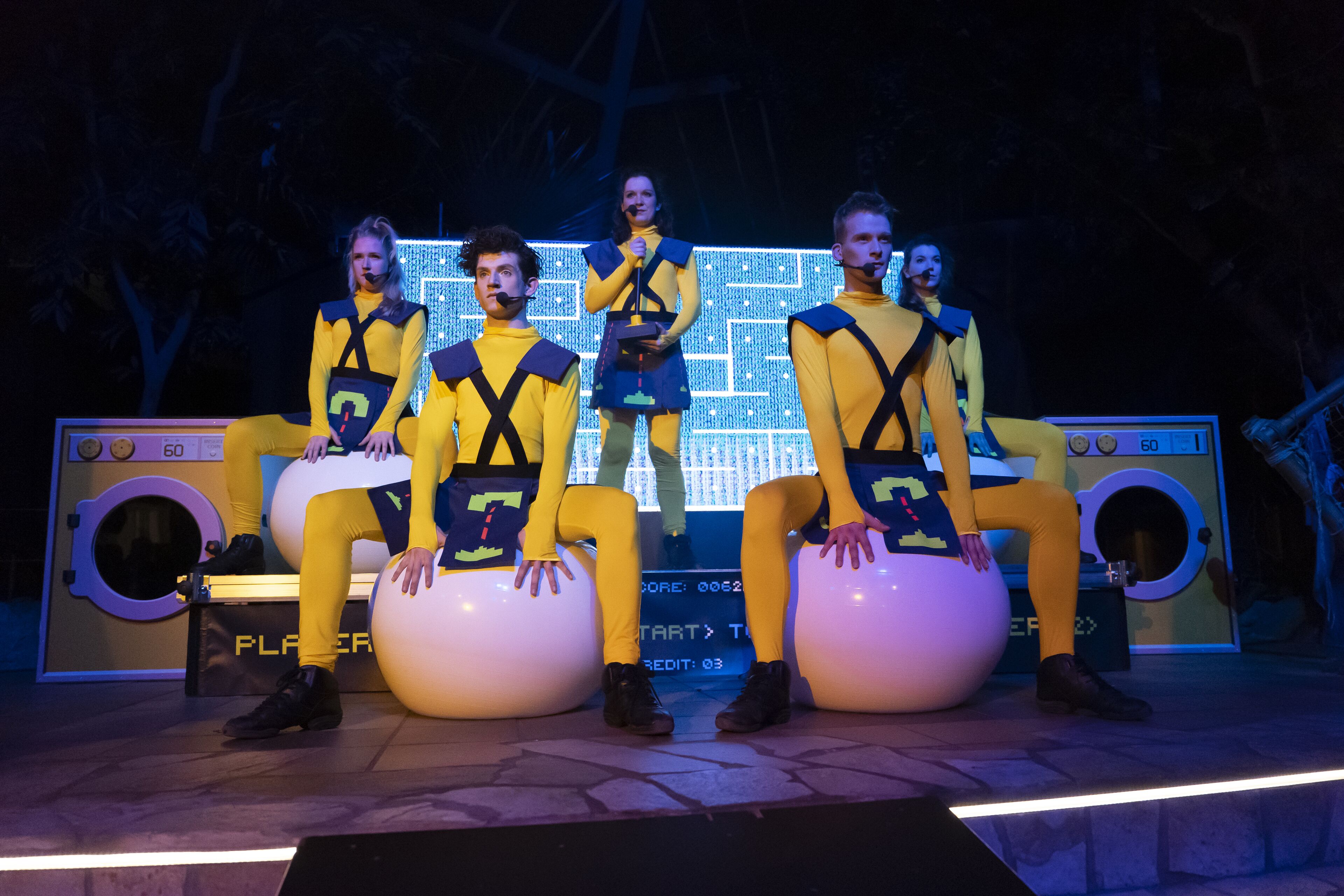 Une scène où quatre personnes, vêtues de jaune, sont assises sur de grandes boules blanches, avec un grand écran illuminé en arrière-plan.