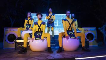 A stage with four people in yellow outfits sitting on large white balls, with a large illuminated screen in the background.