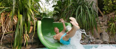 Eine Person saust einen grünen Wasserrutschen in ein Schwimmbecken, das von viel Grün und Felsen umgeben ist.