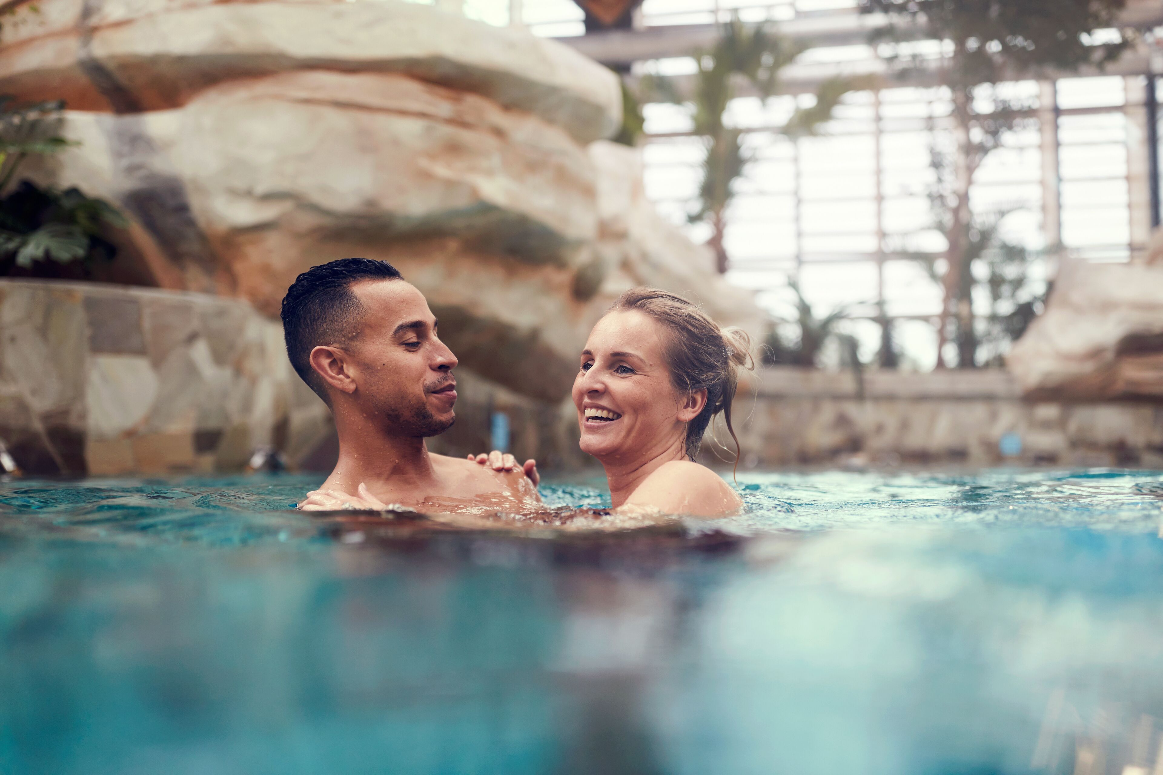 A couple in a swimming pool with a natural rock wall and indoor plants in the background.