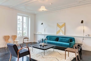 A modern living room with a teal sofa, a coffee table, and two chairs. There is a large window with blinds, a floor lamp, and a painting on the wall.
