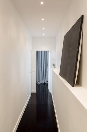 Um corredor estreito com paredes brancas e chão preto, que leva a um quarto com uma pintura grande na parede.