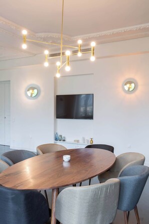 Uma sala de jantar moderna com uma mesa de madeira redonda, seis cadeiras estofadas, um candelabro com várias lâmpadas, uma TV de tela plana fixada na parede e vasos decorativos em uma estante.