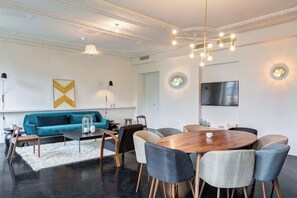 A modern living room with a teal sofa, a round wooden dining table, and a TV mounted on the wall.