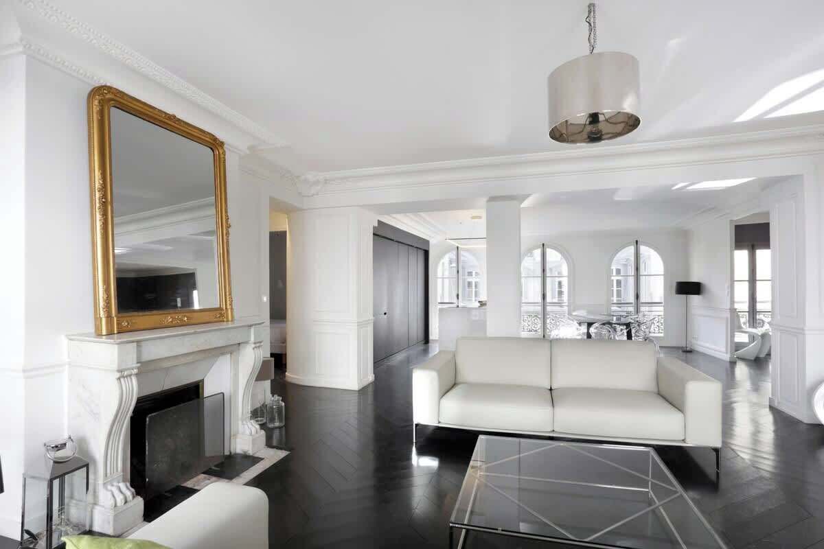 A spacious living room with a white fireplace, a large mirror, a white sofa, and a glass coffee table.