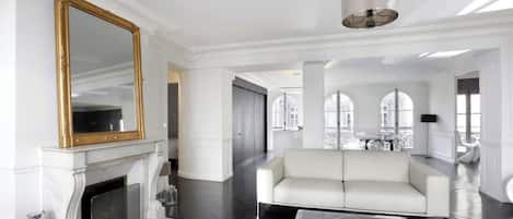 Un salon spacieux avec une cheminée blanche, un grand miroir, un canapé blanc et une table basse en verre.