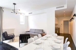 Uma zona de refeições moderna com uma mesa branca posta para seis pessoas, um sofá cinzento e uma cadeira preta.