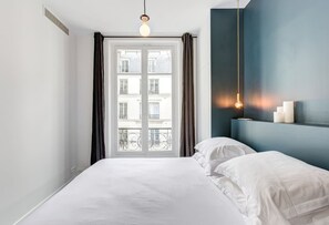Quarto com cama grande, janela com cortinas, parede azul e luminária pendente.