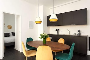 Uma área de refeições moderna com uma mesa de madeira redonda, cadeiras amarelas e azul-turquesa, e luminárias pendentes.