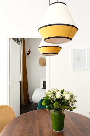 Uma área de refeições com uma mesa de madeira, um vaso verde com flores e uma luminária pendente amarela e branca.