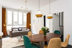 Uma sala de estar moderna com um sofá, mesa de centro e mesa de jantar com cadeiras. Há janelas amplas com cortinas, um lustre e vista para a cidade.