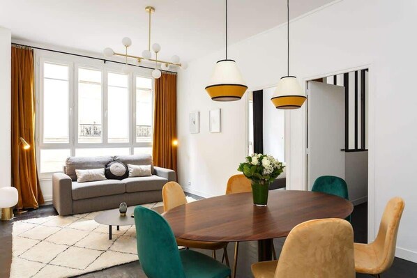 A modern living room with a sofa, coffee table, and dining table with chairs. There are large windows with curtains, a chandelier, and a view of the city.