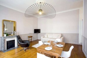A modern living room with a fireplace, a dining table set for four, a white sofa, and a large ceiling light fixture.