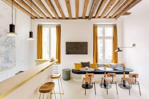 Ein modernes Zimmer mit Holzbalkendecke, einer Bar mit Hockern, einer Sitzecke mit runden Tischen und Stühlen sowie großen Fenstern mit Vorhängen.