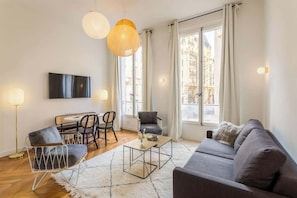 A modern living room with a sofa, chairs, a coffee table, and a TV mounted on the wall. Large windows allow natural light and views of the city.